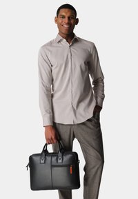 Black leather briefcase with two handles, front zip pocket with orange accent, held by person wearing a light gray button-up shirt and gray trousers.