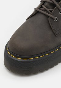 Brun läderankelboots med rund tå, tjock svart gummisula och gula sömmar längs kanten. Strukturera yta med snören.