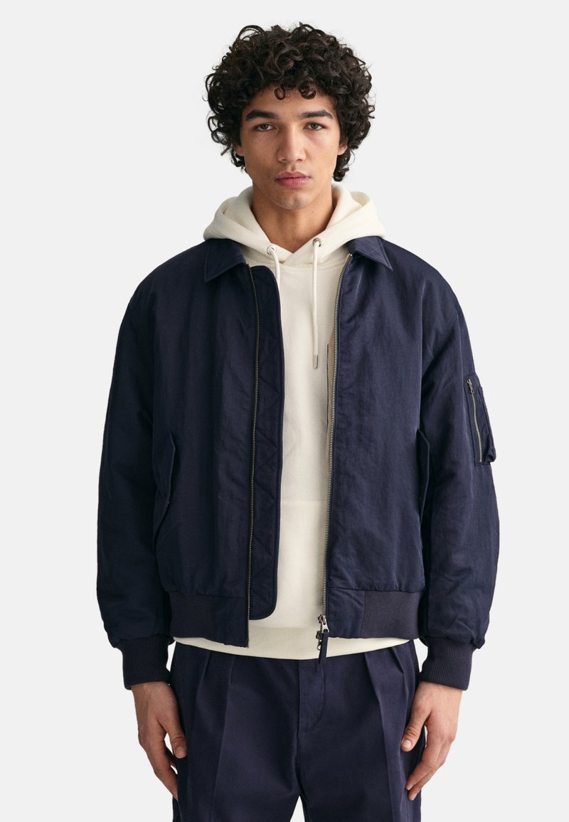 Marineblauer Bomberjacke mit gerippten Bündchen und Saum, vorne mit Reißverschluss und seitlichen Taschen. Getragen über einem weißen Hoodie, der eine Reißverschlusstasche am Ärmel hat.