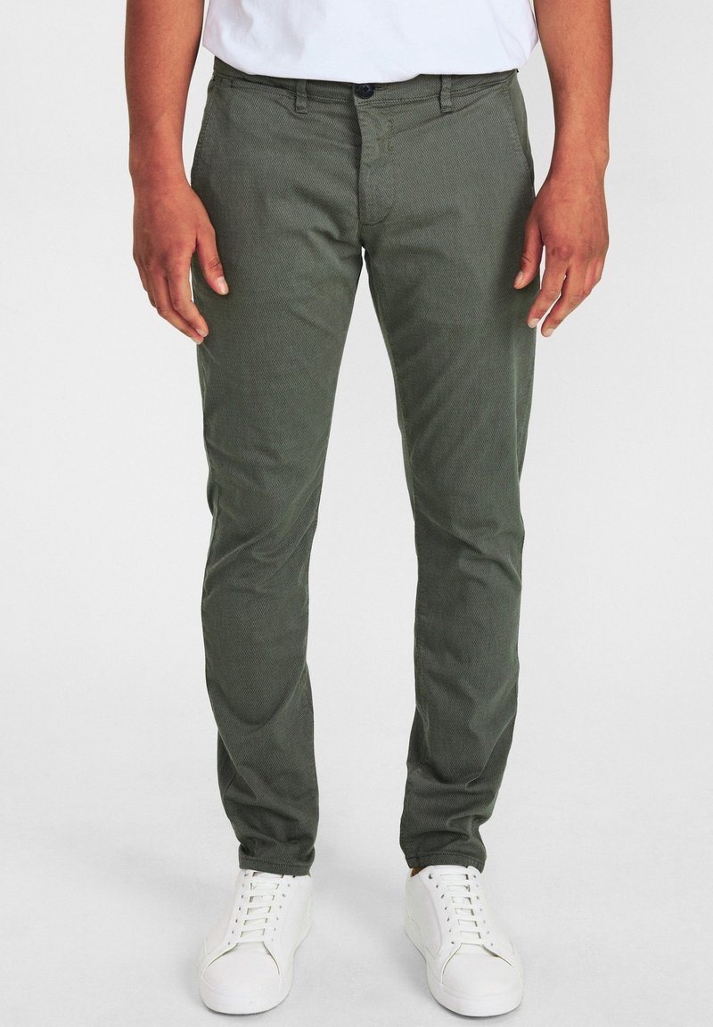 Man som bär slim-fit olivgröna byxor och vita sneakers, står framför en neutral vit bakgrund.