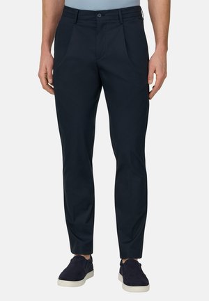 Mężczyzna w dopasowanych ciemnogranatowych spodniach i pasujących granatowych butach wsuwanych, stojący na tle jednolitego jasnego tła.