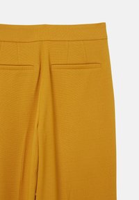 Vista posteriore di pantaloni giallo senape su misura con cintura e due tasche orizzontali su uno sfondo semplice.