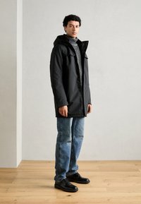 Abrigo exterior negro con capucha, cierre de botones y bolsillos frontales, superpuesto a un cuello de tortuga gris texturizado. Combinado con jeans de denim azul y zapatos negros.