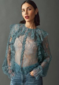 Blouse en dentelle teal avec des manches volantées, un col rond et des détails de boutons en tissu transparent, associée à un bas en denim.