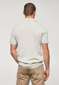 Homme portant un polo à manches courtes gris clair et un pantalon beige, se tenant de dos contre un fond uni clair.
