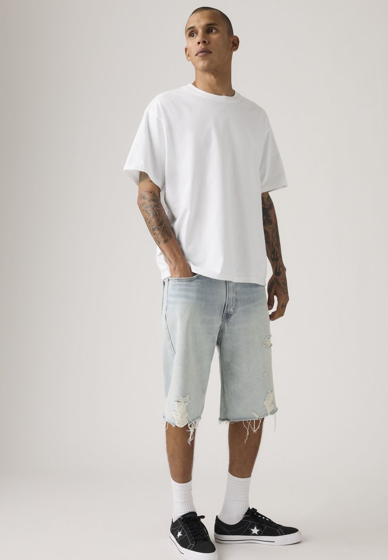 Man met tatoeages, gekleed in een los wit T-shirt, gescheurde lichte denim shorts, witte sokken en zwarte lage sneakers, staand met handen in de zakken.