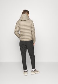 Light beige padded jacket with a hood, featuring horizontal stitching. Worn with dark trousers and cream sneakers, viewed from the back.