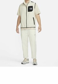 Chaleco de cremallera beige claro con detalles negros y un bolsillo en el pecho, combinado con pantalones beige a juego y zapatillas gris claro.