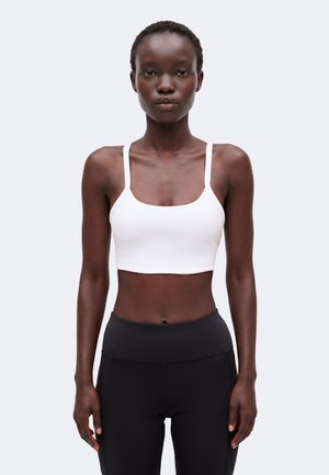 Schlanke Person mit kurzen Haaren, die ein weißes Crop-Top und schwarze Leggings mit hoher Taille trägt, steht vor einem schlichten hellen Hintergrund.