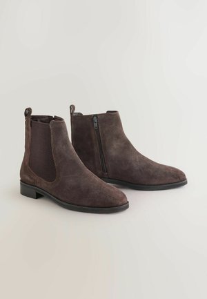 Bottines en daim marron avec un bout pointu, un mesh latéral et un zip, semelle en caoutchouc noir plat, et des tirants à l'arrière pour un enfilage facile.