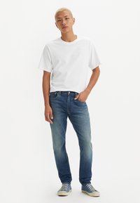 Camiseta blanca de algodón, pantalones vaqueros de denim azul con un acabado desgastado y zapatillas azules. Diseño simple y casual con un corte relajado.