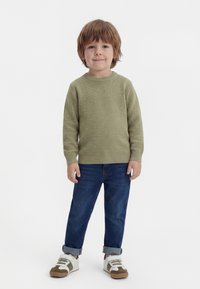 Jongen met bruin haar die een groene trui draagt, opgerolde blauwe spijkerbroek en witte sneakers, staand en glimlachend.