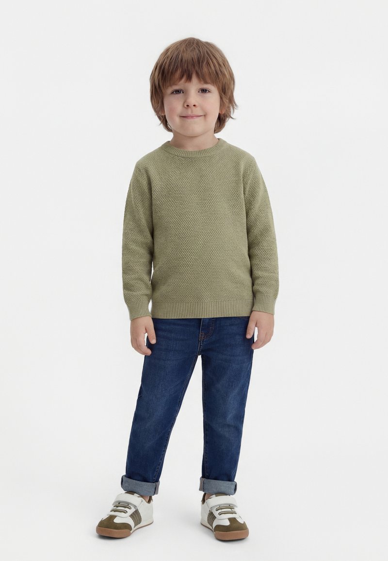 Jongen met bruin haar die een groene trui draagt, opgerolde blauwe spijkerbroek en witte sneakers, staand en glimlachend.