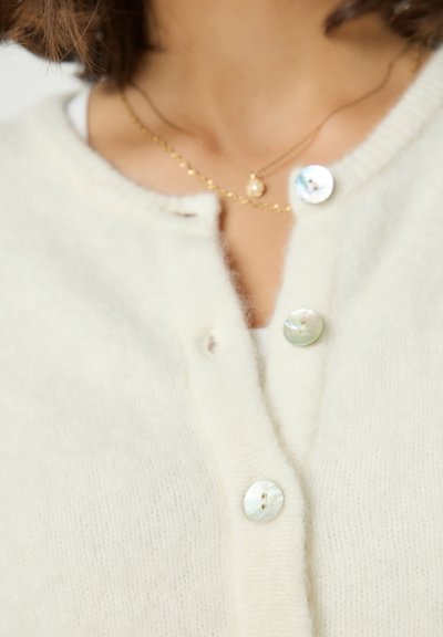 Gros plan sur une personne portant un cardigan en laine crème avec trois boutons nacrés et deux délicats colliers en or.