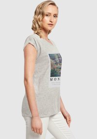 T-shirt gris à manches courtes en tissu doux, arborant un panneau d'œuvre imprimé aux tons verts et bleus. Texte blanc en bas du design.