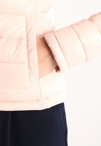 Hellrosa gesteppte Steppjacke mit horizontalem Steppmuster, seitlicher Tasche und enganliegenden Bündchen. Glatte, leicht glänzende Textur.