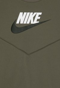 Olīvu zaļš Nike džemperis ar balto logo uz krūškurvja un melnu „swoosh”. Gluda tekstūra ar kontrastējošiem steidzējumiem.