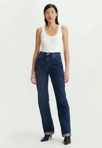 Jeans dritti blu scuro in denim, con vita alta, design a cinque tasche e un leggero sfilacciamento all'orlo. Abbinati a un top bianco senza maniche.