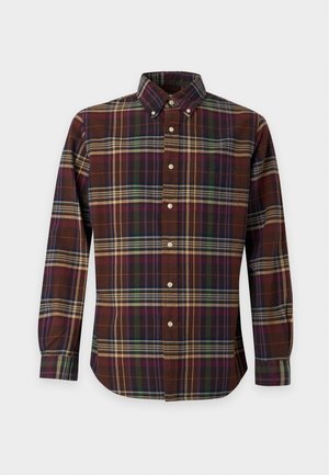 Jeune homme debout portant une chemise à carreaux foncée boutonnée, un jean bleu clair ample et des mocassins beiges sur un fond blanc uni.