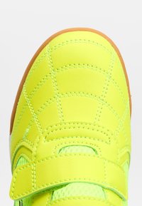 Gros plan sur l’avant d’une chaussure jaune néon avec des coutures détaillées et un bord de semelle marron, avec une bride Velcro sur un tissu respirant.