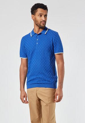 Blauwe, textuurvolle polo met korte mouwen, witte kraag en accenten op de mouwen, gecombineerd met beige broeken. Detail met drie knopen bij de hals.