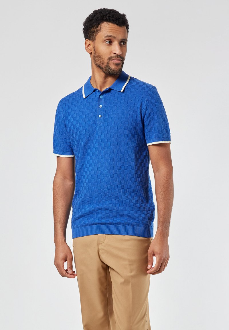 Sininen tekstuurinen polo-paita lyhyillä hihoilla, valkoisella kauluksella ja hihojen korostuksilla, yhdistettynä beigeihin housuihin. Kolmen napin yksityiskohta.