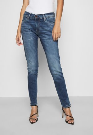 Femme portant un jean skinny délavé moyen et des sandales à talons noirs, debout devant un fond uni gris et blanc.