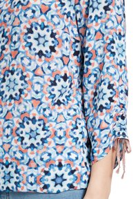 Bunt gemusterte Bluse mit einem floralen kaleidoskopischen Design. Hergestellt aus leichtem Stoff, mit gerafften Ärmeln und dekorativen Bindebändern.