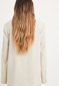 Cream-colored tailored blazer with structured shoulders and long sleeves, viewed from the back, paired with wavy, shoulder-length hair.