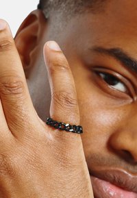 Zwart gevlochten ring met witte accenten, gedragen op een vinger. De ring heeft een textuur oppervlak en een flexibele design, afgebeeld tegen de huid van een persoon.