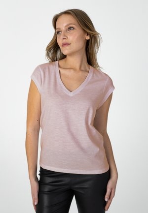 Femme portant un haut rose clair à manches courtes et col en V et un pantalon noir, debout avec la tête légèrement tournée vers la gauche sur un fond blanc.