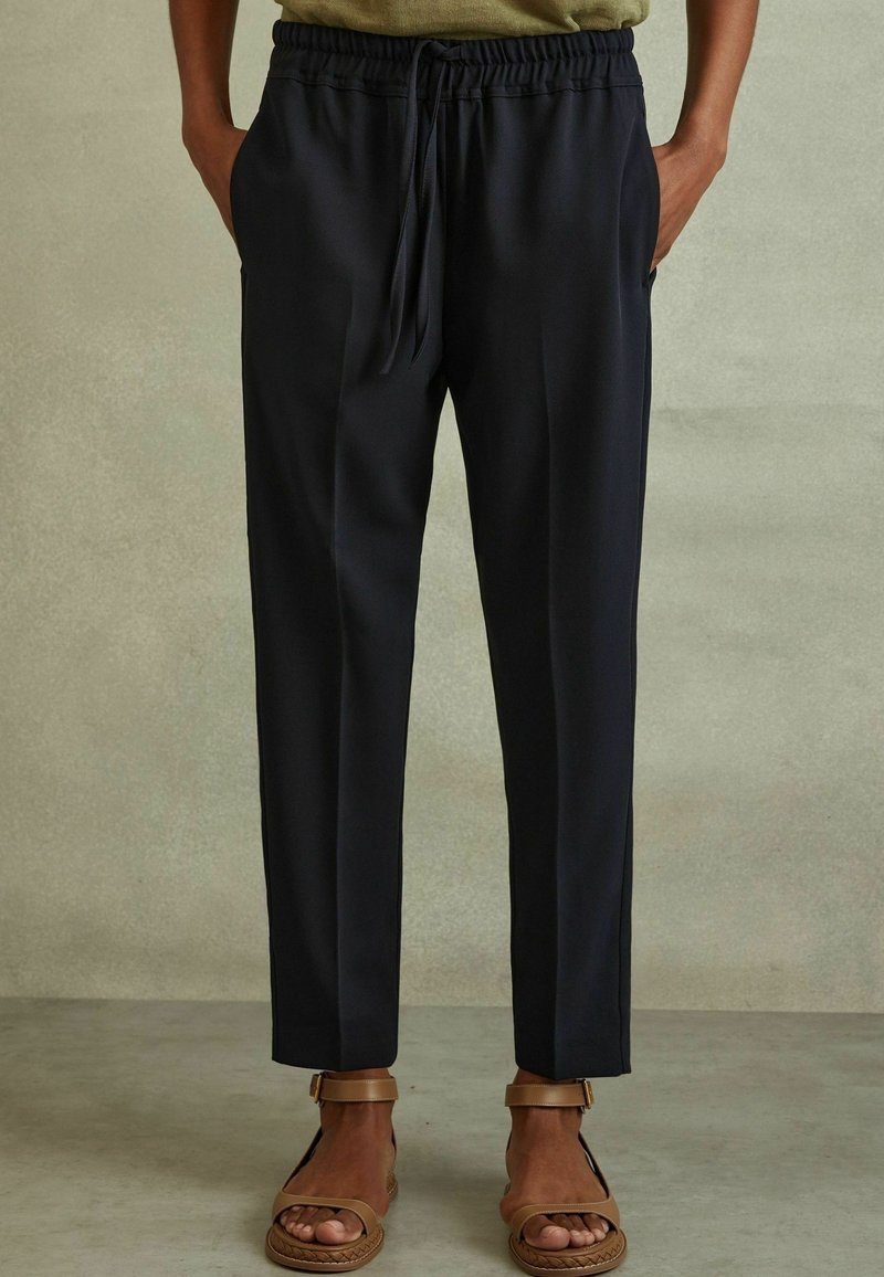 Personne portant un pantalon noir à cordon et des sandales ouvertes marron, debout les mains dans les poches devant un fond uni.