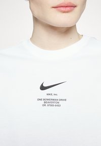 Vit T-shirt med en svart Nike swoosh-logotyp och varumärkesinformation i svart text på framsidan. Mjuk bomullsmaterial, design med rund hals.