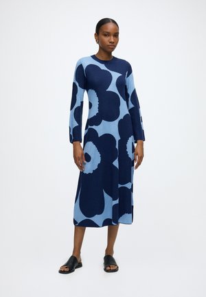 Femme debout portant une robe bleue midi à manches longues avec de grands motifs floraux bleu foncé et des sandales noires ouvertes à l’avant.