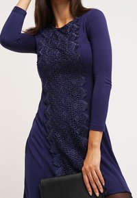 Mörkblå långärmad klänning med texturerade cirkulära spetsdetaljer längs fram, tillsammans med en svart kuvertväska. Slät stickad tyg.