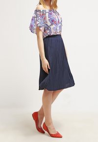 Blusa off-the-shoulder con motivo floreale colorato, abbinata a una gonna plissettata blu navy. Completa l'outfit un paio di scarpe rosse con dettagli a intaglio.