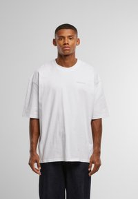 Bílé bavlněné tričko s volným střihem, kulatým výstřihem, krátkými rukávy a jemným logem v světlejší barvě. Jednoduchý povrch, neformální design.