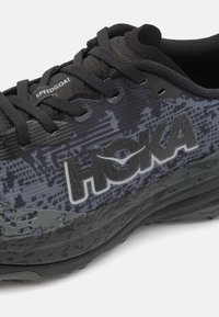 Zapatilla de running en negro y gris con parte superior texturizada, suela gruesa y logo "HOKA". Presenta un diseño con patrón y cordones negros.