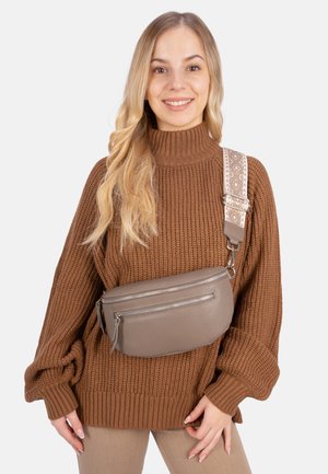Brauner gestrickter Pullover über hellbeigen Hosen mit einer hellbraunen Lederbauchtasche, die zwei Reißverschlüsse und einen gemusterten verstellbaren Riemen hat.