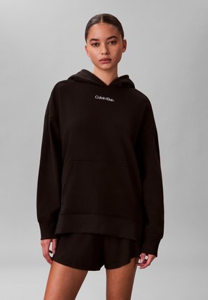 Junge Frau mit zurückgebundenem Haar, die einen schwarzen Calvin Klein Hoodie und passende schwarze Shorts vor einem hellgrauen Hintergrund trägt.
