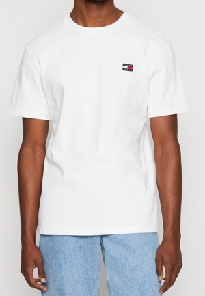 Mężczyzna ubrany w gładką białą koszulkę z krótkim rękawem z małym logo Tommy Jeans na klatce piersiowej, w połączeniu z jasnoniebieskimi jeansami.