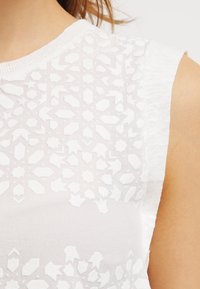 Vit ärmlös topp med ett texturerat mönster av geometriska former. Mjuk tyg med en stickad kant vid halsringningen och axelområdet.