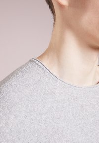 Hellgrauer Pullover aus einem weichen, strukturierten Stoff. Hat einen runden Ausschnitt und sichtbare Nähte am Rand. Nahaufnahme des Schulterbereichs.