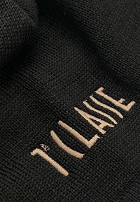 Tessuto nero lavorato a maglia con testo ricamato beige che recita "I'CLAIRE." La trama è liscia con un'intrecciatura fitta e fibre libere minime.