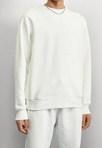 Man i off-white sweatshirt och matchande sweatpants med silver kedja, står mot en enfärgad ljus bakgrund.