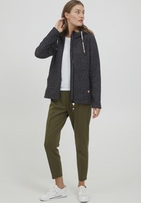Mörkgrå zip-up jacka med huva och trädetaljer, kombinerad med olivgröna joggingbyxor och vita sneakers. Modellen visas mot en neutral bakgrund.
