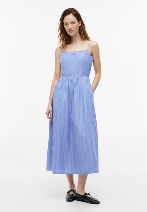 Γυναίκα που στέκεται φορώντας ένα μίντι φόρεμα με κάθετες μπλε και λευκές ρίγες χωρίς μανίκια, με τσέπες και μαύρα επίπεδα παπούτσια, μπροστά σε απλό φόντο.