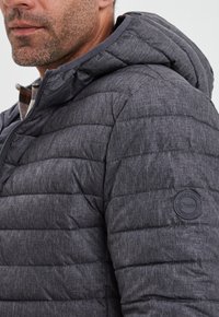 Giacca grigia imbottita con una finitura testurizzata, dotata di cappuccio e una patch rotonda con logo sulla manica. Dettaglio di trapuntatura orizzontale su tutta la giacca.