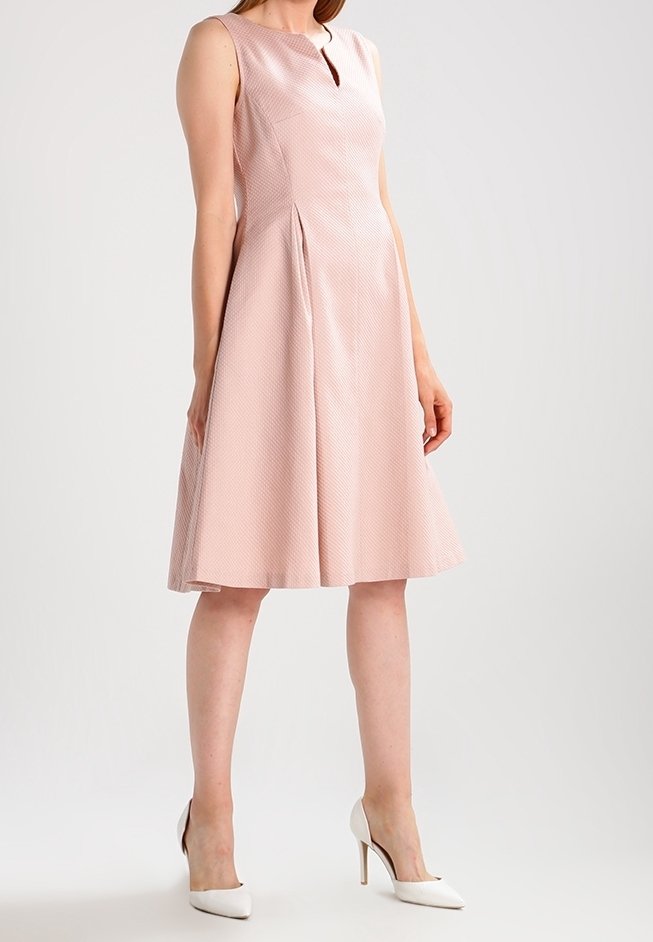Femme portant une robe rose clair sans manches, longueur genou, avec des plis, associée à des chaussures blanches à talons hauts sur un fond uni.
