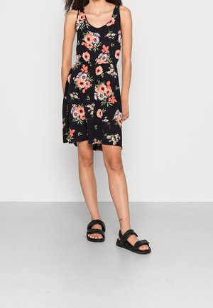 Vrouw draagt een zwarte mouwloze jurk met roze en witte bloemenprint en zwarte sandalen met open tenen, staand tegen een effen lichte muur.